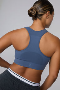 On a light gray studio backdrop, India wears the Seamless Racerback Sports Bra in Evernight with charcoal joggers. The back view showcases the racerback design, breathable textured knit panels, wide underband, and tonal Harper Wilde logo centered on the band.