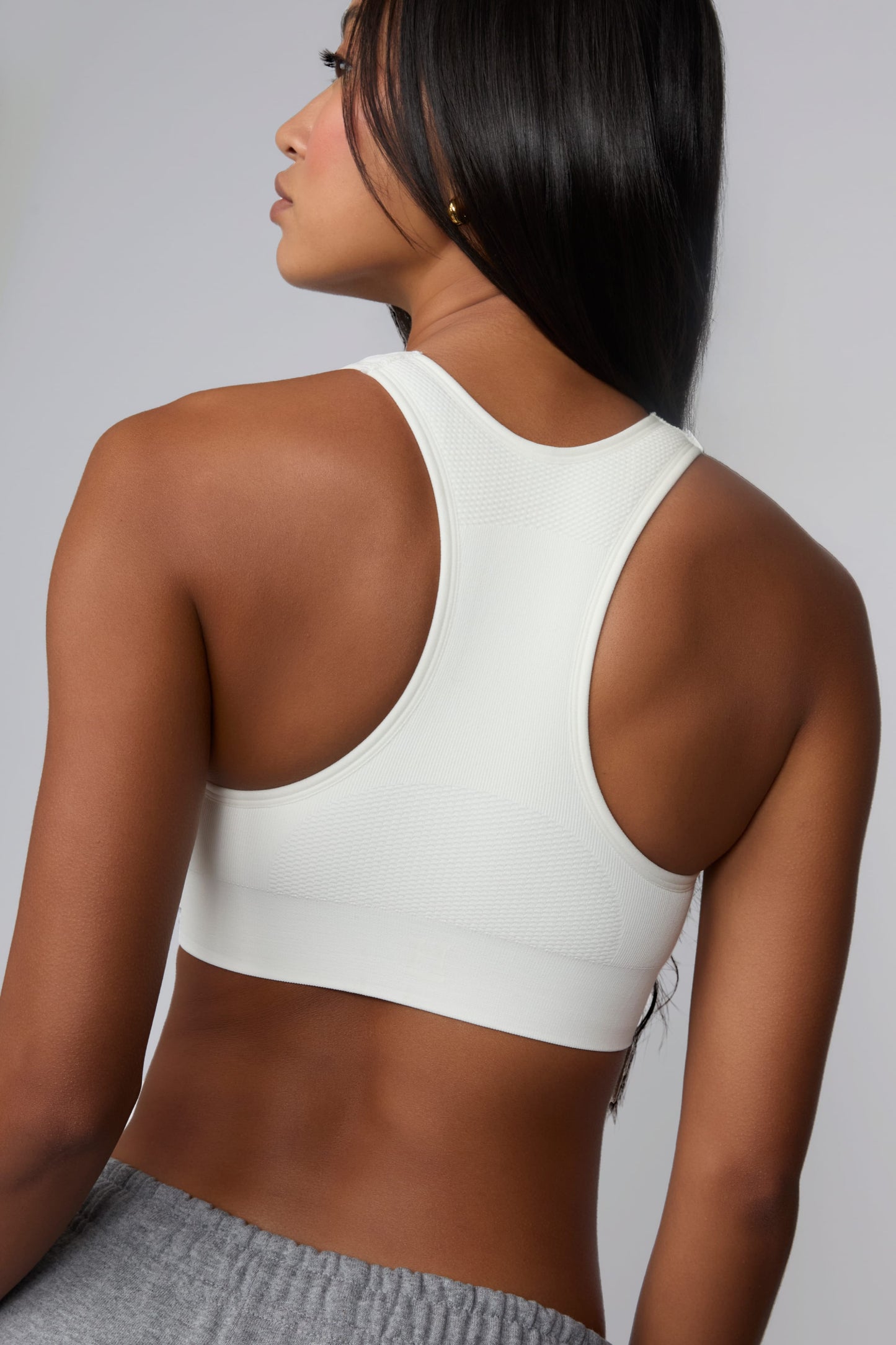 On a light gray studio backdrop, Amber wears the Seamless Racerback Sports Bra in White with gray sweatpants. The angled back view highlights the racerback silhouette, textured knit details, and wide underband.