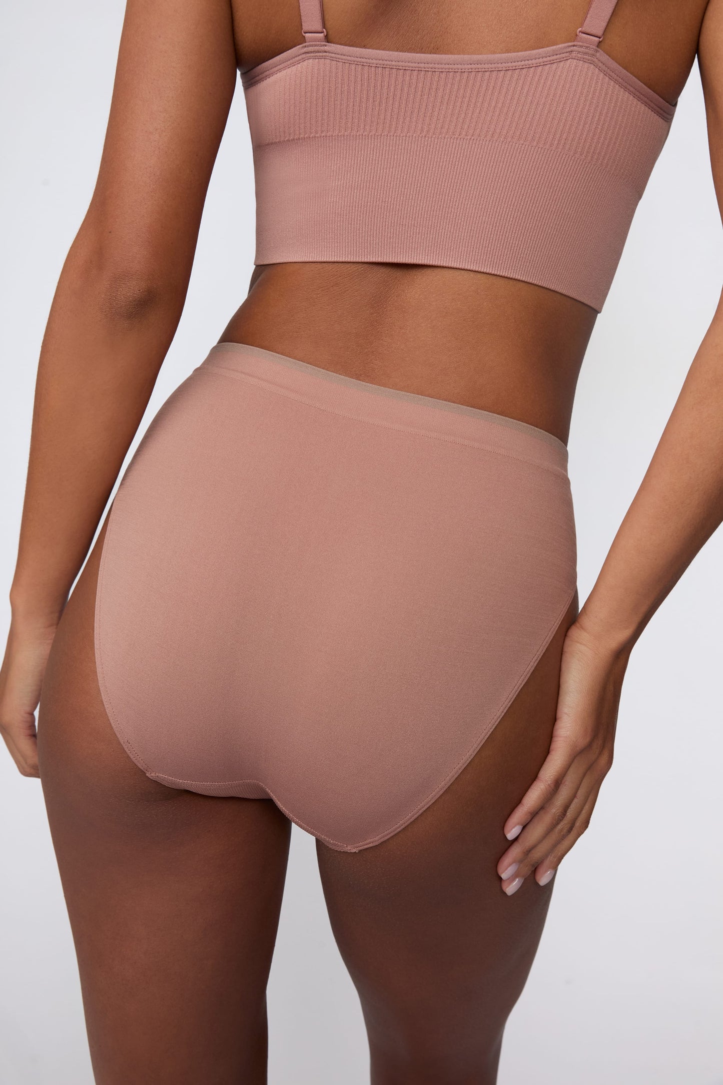 In a studio setting, Courtney wears the Smooth Seamless Brief in Plum with the matching Seamless Scoop Bralette in Plum. The back view highlights the full back coverage, smooth finish, and contrast trim waistband.
