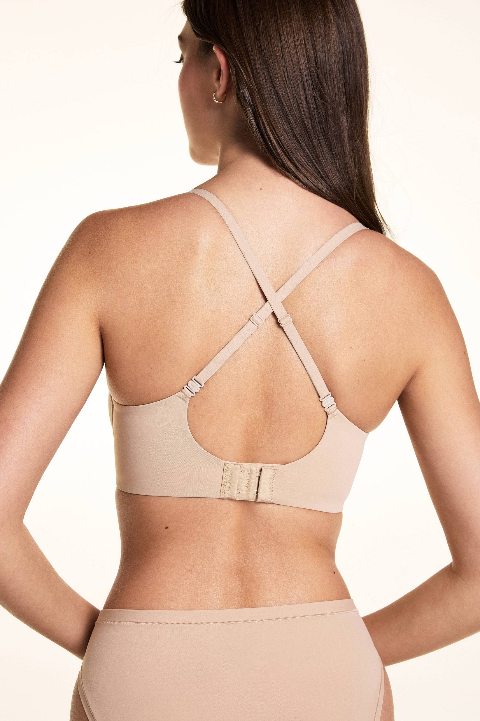 On a light neutral backdrop, Laura is shown from the back wearing the Base Balconette Bra in Beige with the straps crossed. The image highlights the convertible crisscross straps, wide back band, and hook-and-eye closure.
