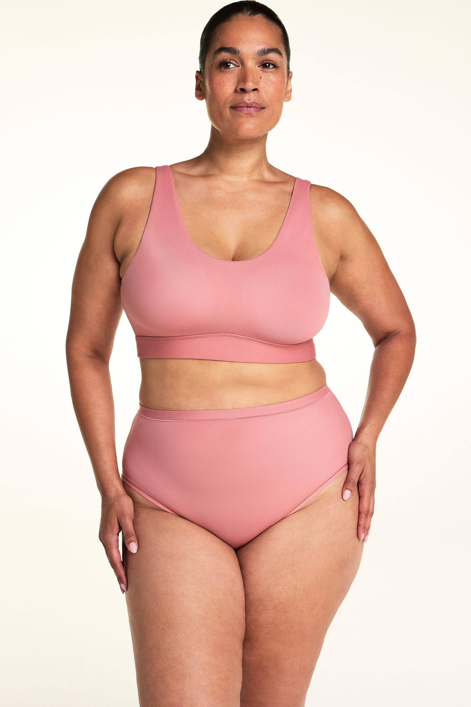 On a light neutral backdrop, Jennifer wears the Bliss Scoop Adjustable Bralette in Cinnamon with matching high-rise underwear. She stands facing forward, showing the scoop neckline, wide straps, and smooth wireless design.