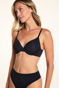 On a light studio backdrop, Meagan wears the Base Picot T-Shirt Bra in Black in a three-quarter view. The image highlights the smooth molded cups, picot trim along the neckline, and adjustable straps.