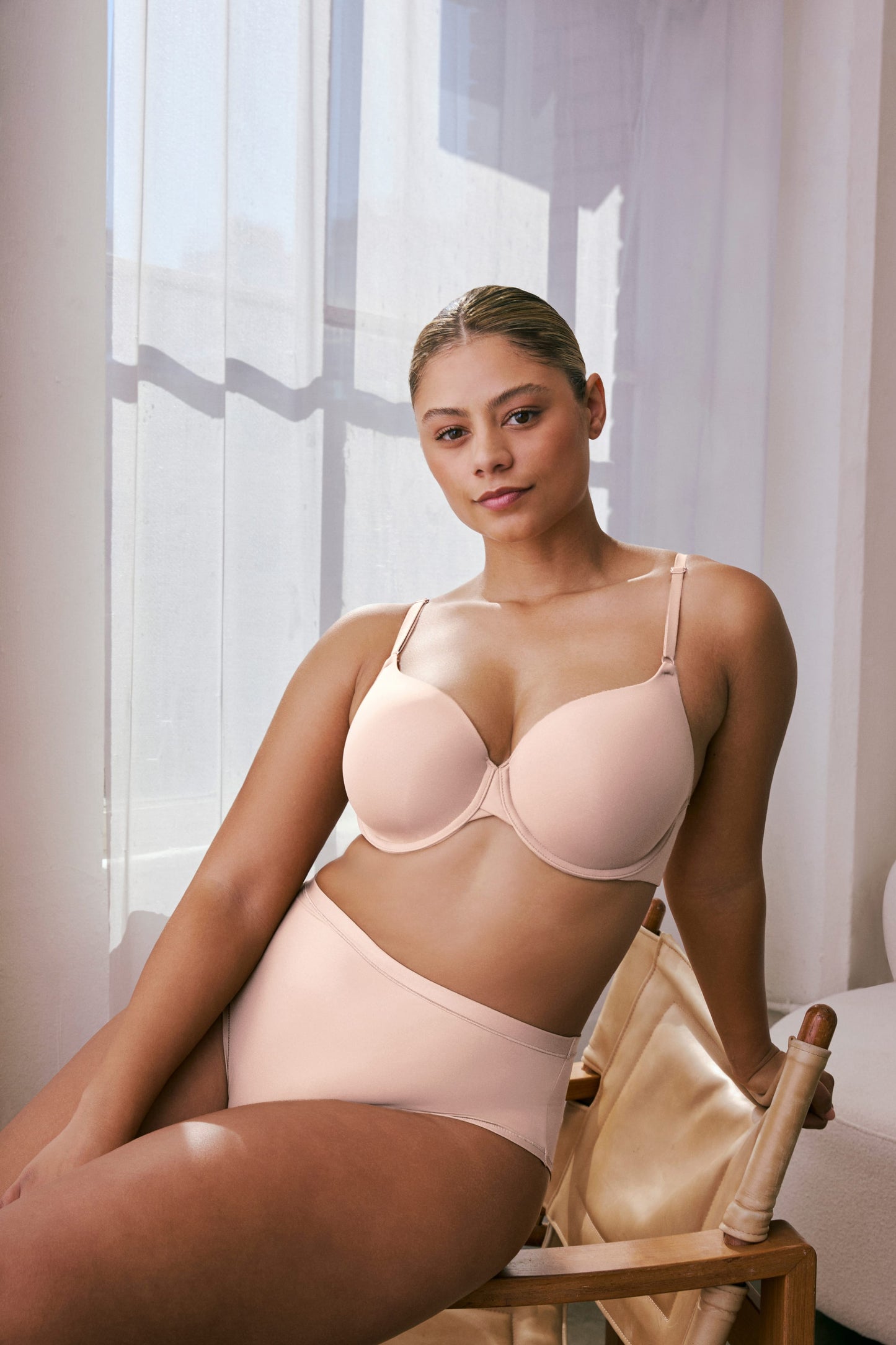 In a sunlit room, India wears the Base T-Shirt Bra in Rose with matching bottoms, seated on a chair. The bra features smooth molded cups, adjustable straps, and an underwire for support. sizing: India wears size 36D