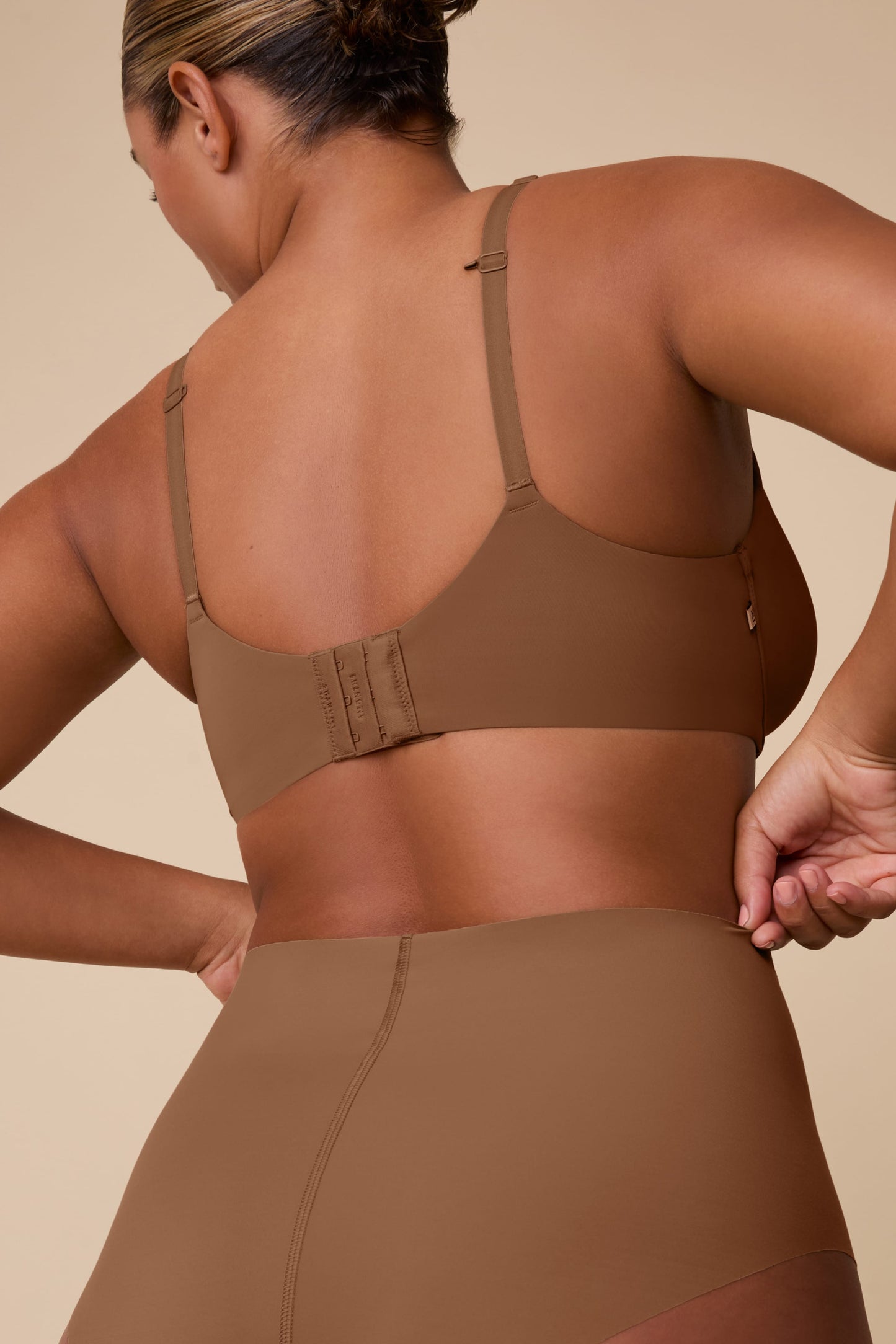 On a neutral beige backdrop, India wears the Base Unlined Bra in Brown, shown from the back to highlight the smooth band, adjustable straps, and hook-and-eye closure.
