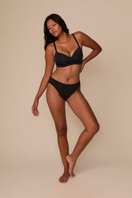 On a neutral beige backdrop, Victoria wears the Base Wirefree T-Shirt Bra in Black with matching underwear, showcasing the smooth cups, wireless support, and adjustable straps in a full-body view.