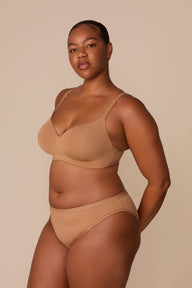 On a neutral beige backdrop, Zarria wears the Base Wirefree T-Shirt Bra in Tan with matching underwear, showcasing the smooth molded cups, wireless support, and adjustable straps from an angled view.