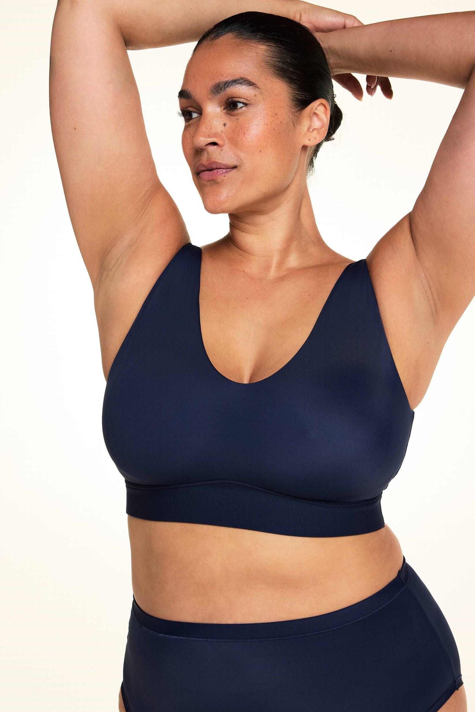 On a light studio backdrop, Jennifer wears the Bliss Bralette in Navy with matching underwear. With both arms raised overhead, she shows the V-neck silhouette, wireless band, and smooth microfiber construction.