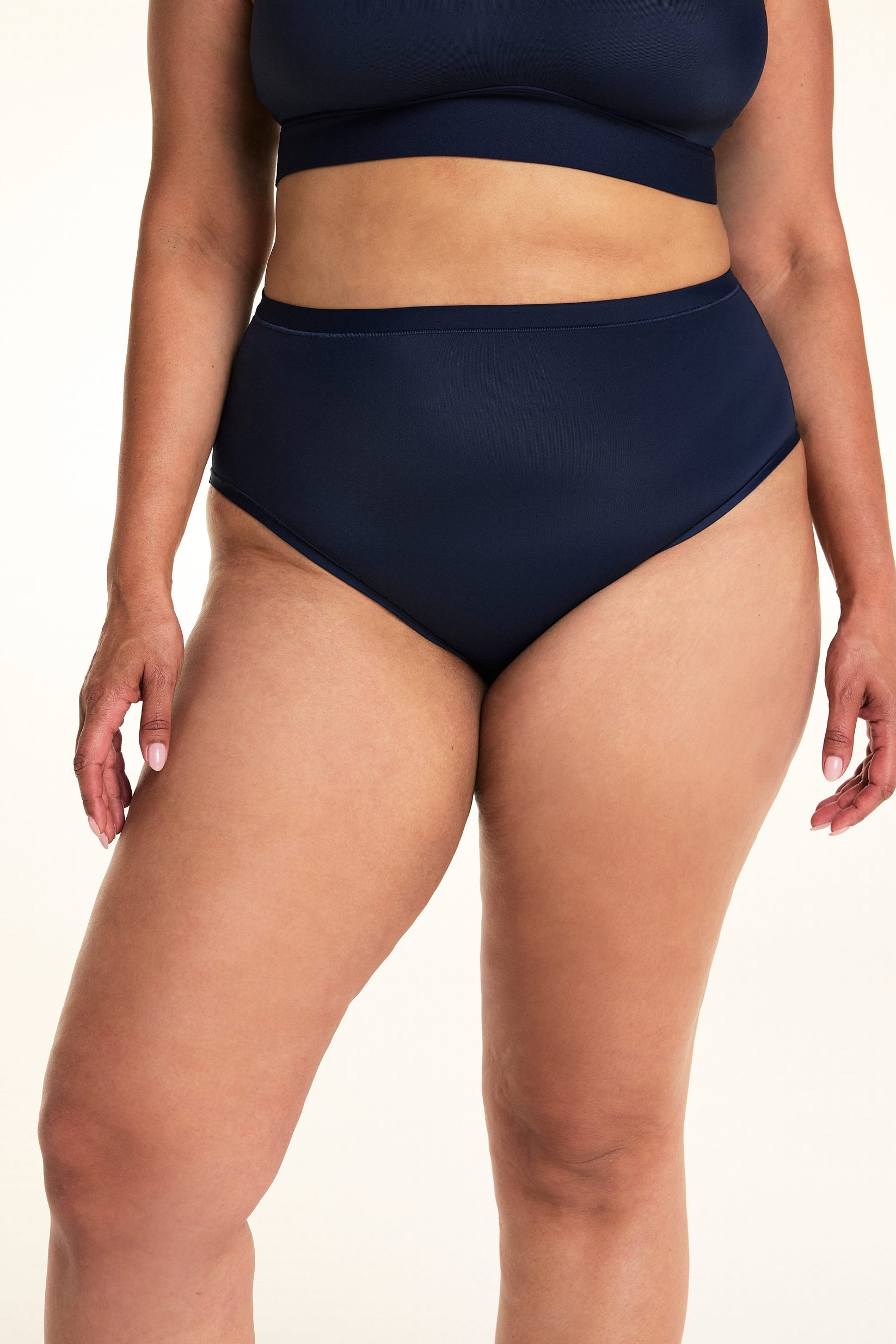 On a light studio backdrop, Jennifer wears the Bliss Highwaist in Navy. The close-up front view highlights the high-rise waistband, smooth fabric, and full-coverage silhouette.