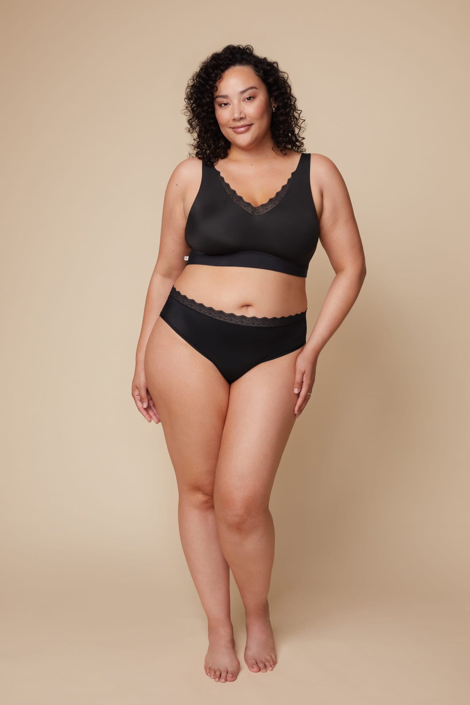 On a neutral beige backdrop, Bianca wears the Bliss Lace Bralette in Black with coordinating underwear, showcasing the deep V-neckline with scalloped lace trim, smooth fabric, and supportive underband in a full-body front view. sizing: Bianca wears size XL