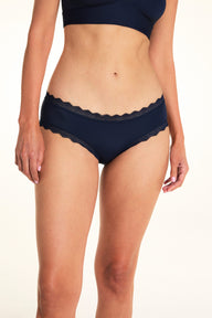 On a light studio backdrop, Meagan wears the Bliss Lace Cheeky in Navy with the Bliss Lace Halterneck Bralette in Navy. The full-body image highlights the smooth fabric, low-rise silhouette, and scalloped lace trim.