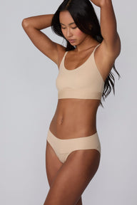 On a light gray studio backdrop, Amber wears the Bliss Low Rise Bikini in Beige with the matching Bliss Bralette in Beige. The angled front view highlights the low-rise waistband and smooth, lightweight design.