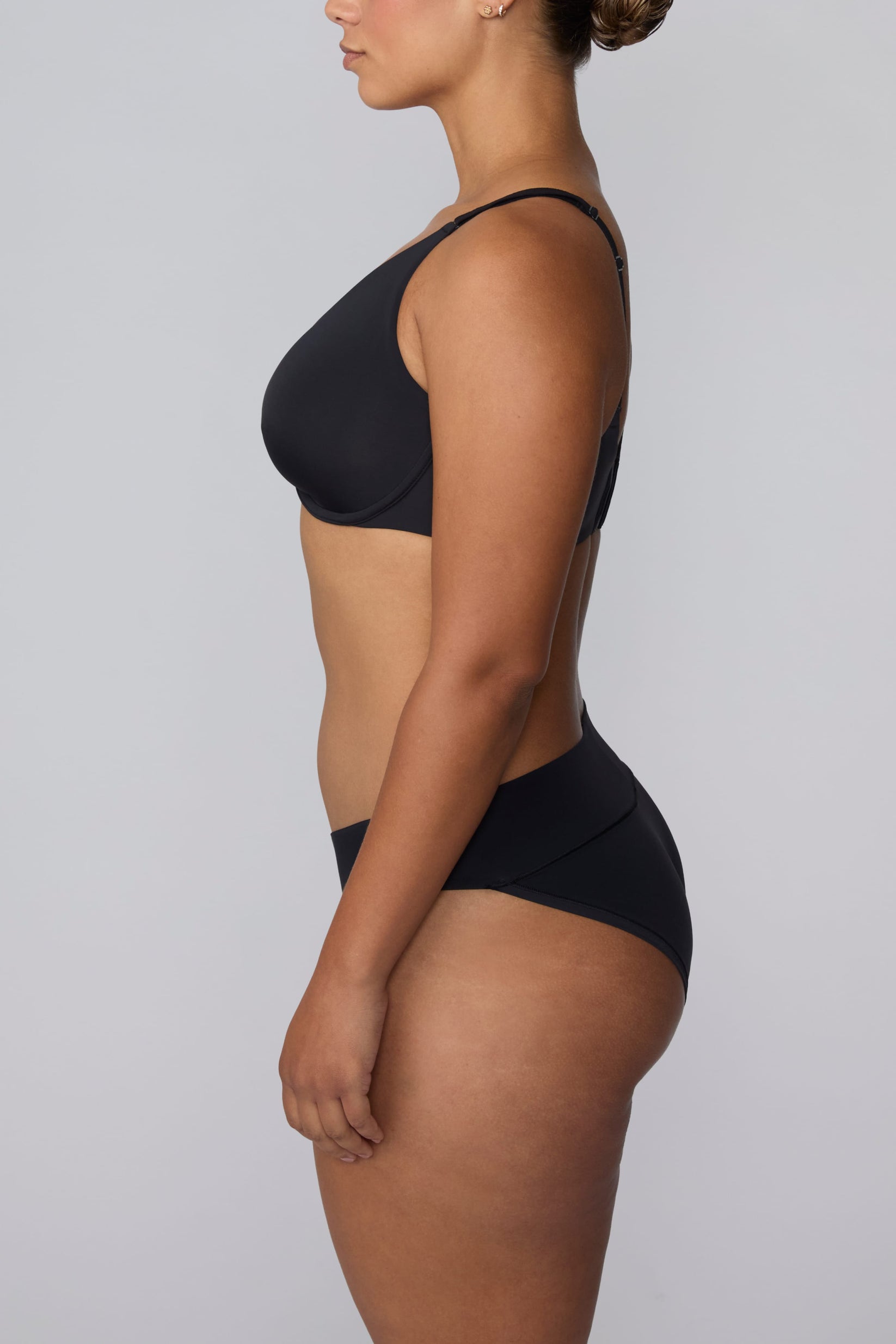 On a light gray studio backdrop, India wears the Bliss Low Rise Bikini in Black with the matching bra in Black. The side profile highlights the low-rise fit and smooth silhouette.
