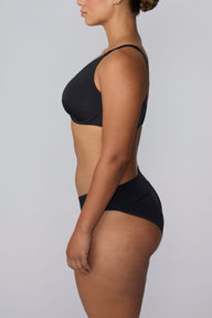On a light gray studio backdrop, India wears the Bliss Low Rise Bikini in Black with the matching bra in Black. The side profile highlights the low-rise fit and smooth silhouette.
