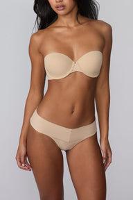 On a light gray studio backdrop, Amber wears the Bliss Low Rise Thong in Beige with the matching Base Strapless Bra in Beige. The front view highlights the smooth waistband, low-rise fit, and lightweight thong silhouette.