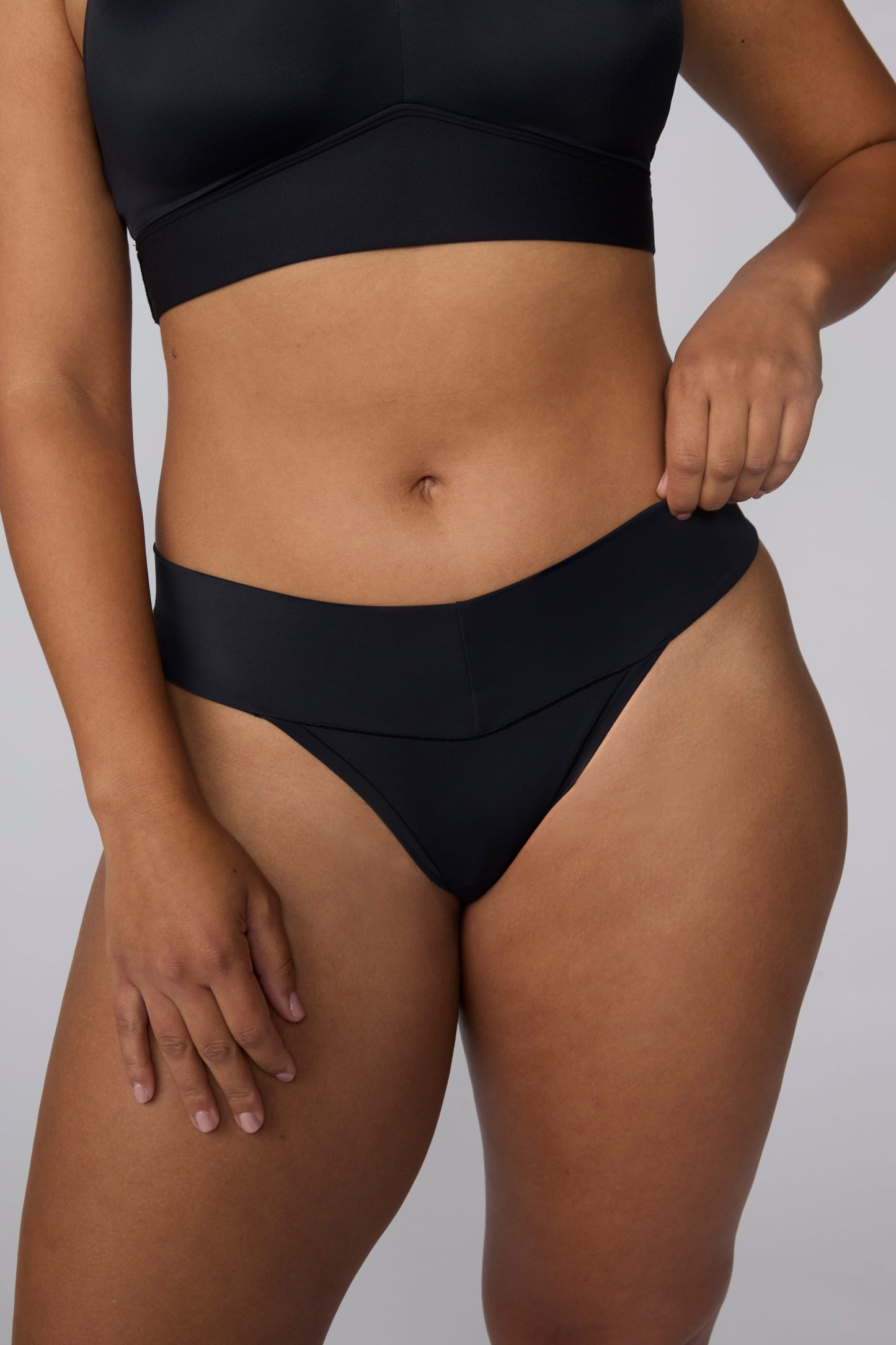 On a light gray studio backdrop, India wears the Bliss Low Rise Thong in Black with the matching Bliss Bralette in Black. The close-up front view highlights the smooth waistband, low-rise fit, and lightweight thong design.