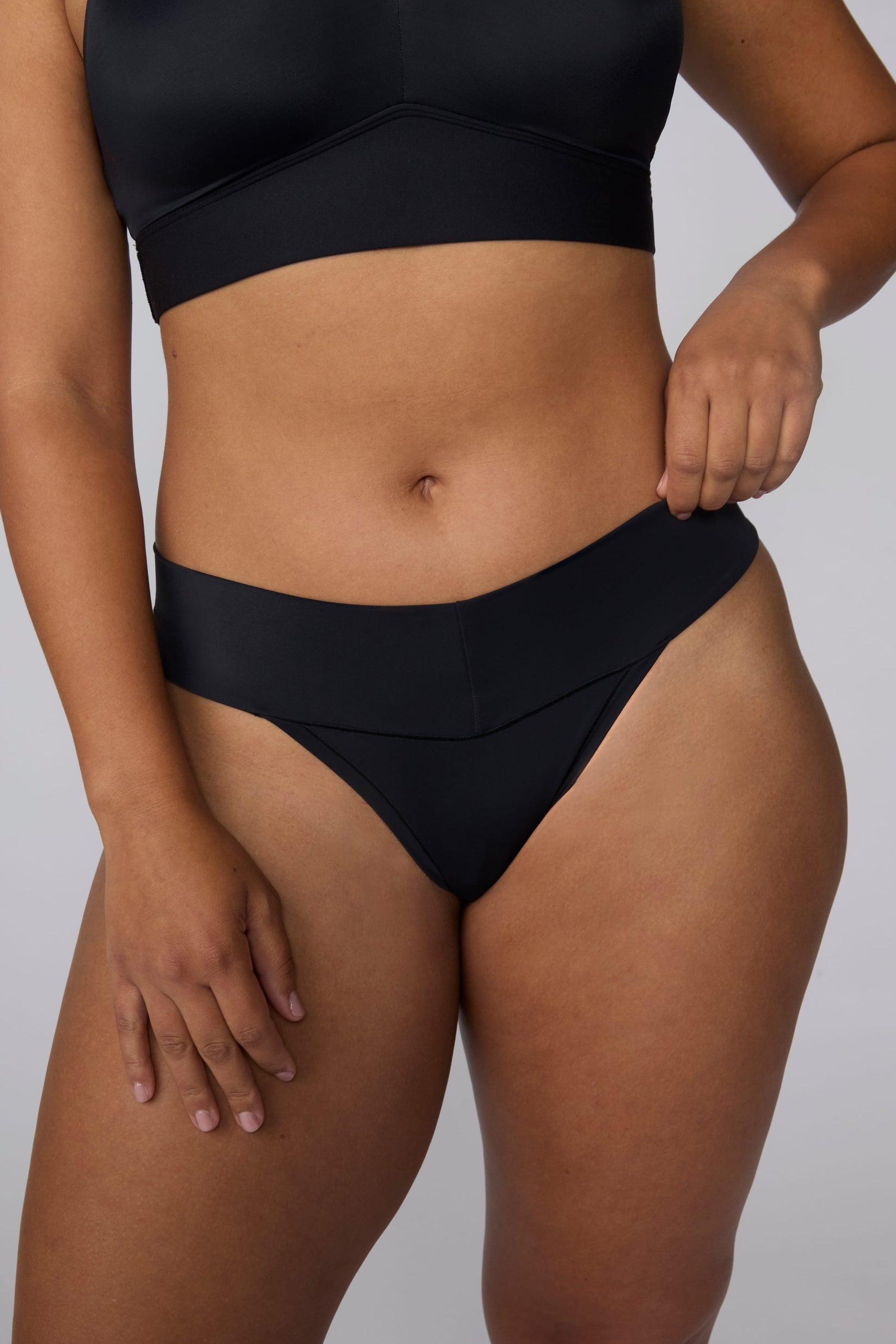 On a light gray studio backdrop, India wears the Bliss Low Rise Thong in Black with the matching Bliss Bralette in Black. The close-up front view highlights the smooth waistband, low-rise fit, and lightweight thong design.