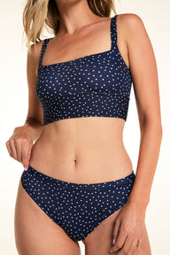On a light studio backdrop, Meagan wears the Bliss Thong in Navy Dot with a matching Navy Dot bralette. The close-up image highlights the low-rise waistband, smooth leg openings, and allover white dot print.