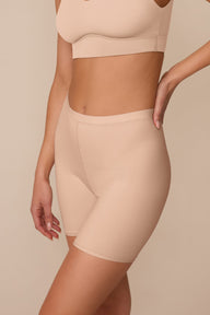 On a neutral studio backdrop, Marcela wears the Bliss Under Short in Beige with a matching beige bralette. The angled side view highlights the smooth fabric, mid-rise fit, and comfortable thigh coverage.