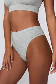 On a light gray studio backdrop, Courtney wears the Cloud Cotton Highwaist Thong in Heather Grey. The angled close-up view showcases the high-rise waistband, high-leg opening, and smooth cotton fabric.
