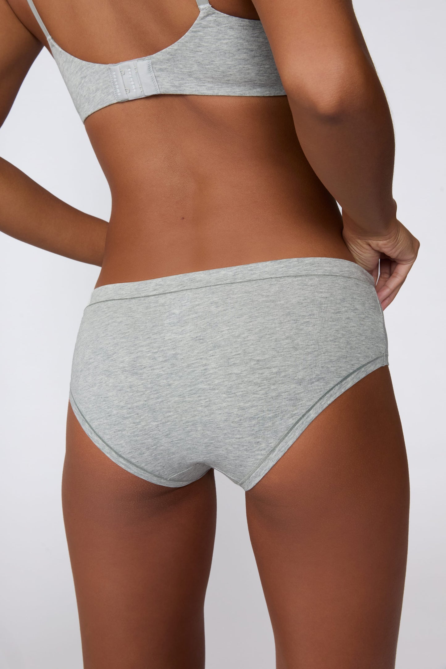 On a light gray studio backdrop, Amber wears the Cloud Cotton Hiphugger in Heather Grey with the matching bra. The angled back view highlights the full back coverage, low-rise waistband, and soft cotton fabric.