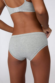 On a light gray studio backdrop, Amber wears the Cloud Cotton Hiphugger in Heather Grey with the matching bra. The angled back view highlights the full back coverage, low-rise waistband, and soft cotton fabric.