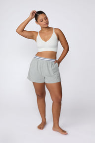 On a light gray studio backdrop, India wears the Cloud Cotton Pajama Short in Heather Grey with a white ribbed bralette. The full-body view highlights the relaxed fit, striped elastic waistband, side pockets, and soft cotton fabric.