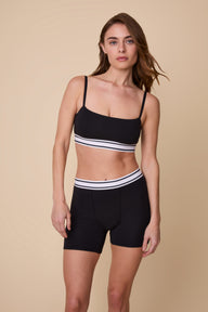On a neutral studio backdrop, Gracie wears the Cloud Cotton Retro Bandeau Bralette and Cloud Cotton Retro Boxer Brief in Black. She stands facing forward, showcasing the bandeau-style square neckline, thin adjustable straps, and matching retro logo waistbands. The soft cotton modal fabric and full-coverage boxer brief design are visible. sizing: Gracie wears size Small