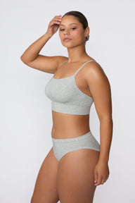 On a light gray studio backdrop, India wears the Cloud Cotton Thin Strap Bralette in Heather Grey with matching underwear. The side profile highlights the straight neckline, thin straps, and soft cotton fabric.