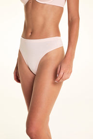 On a light studio backdrop, Alexis wears the Cloud Cotton Highwaist Thong in Dogwood with a matching bra in Dogwood. The angled view highlights the high-rise waistband, high-cut leg opening, and soft cotton fabric. sizing: Alexis wears size Small