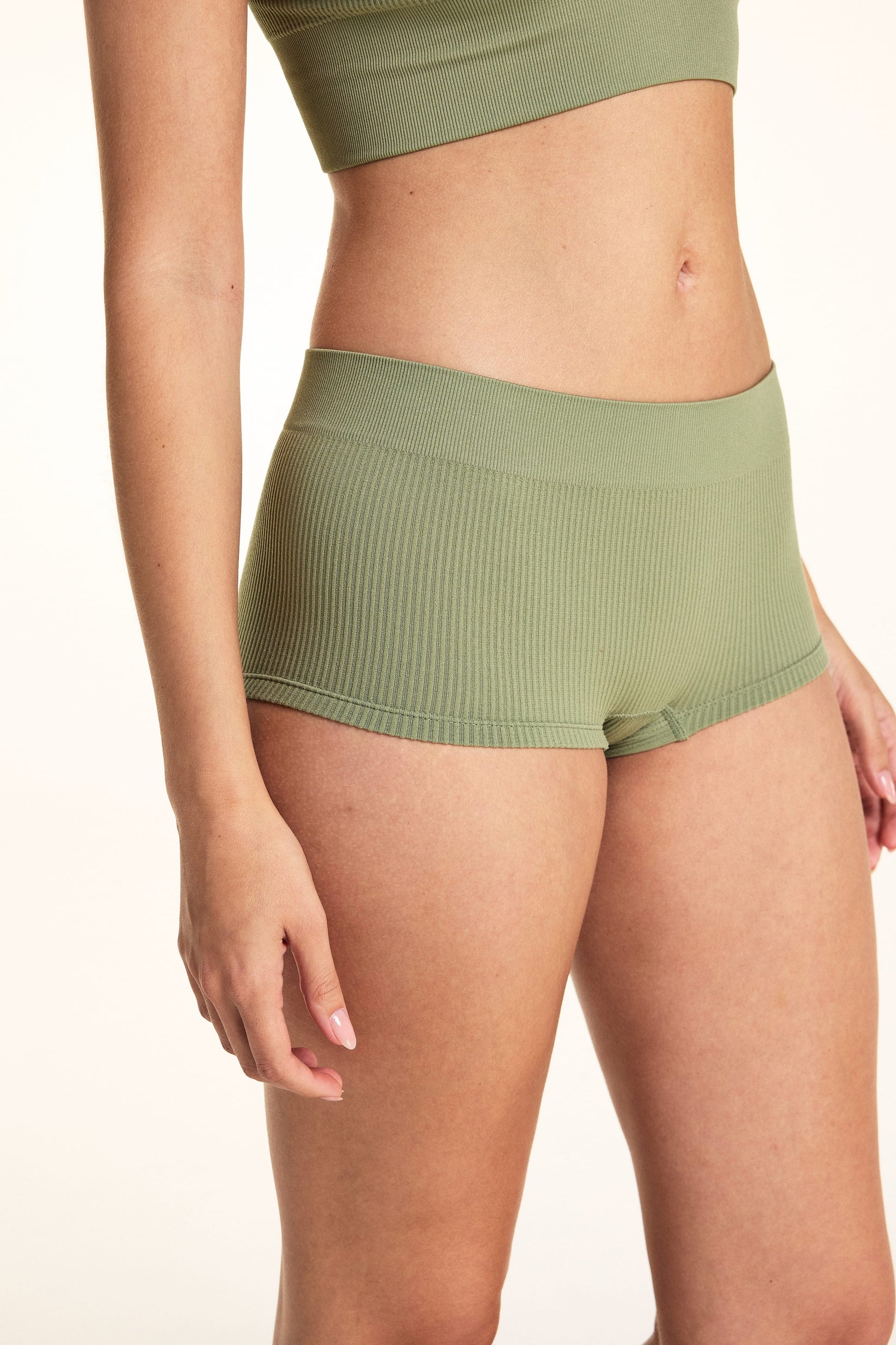 On a white studio backdrop, a side view of Laura wearing the Lounge Rib Sleepshort in Dill highlights the ribbed fabric, seamless construction, and short silhouette.