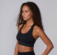 On a neutral studio backdrop, Courtney is shown in a side-front angle wearing the Seamless Racerback Sports Bra in Black with matching bottoms. The smooth scoop neckline, wireless support, and racerback straps are visible as she looks off to the side.