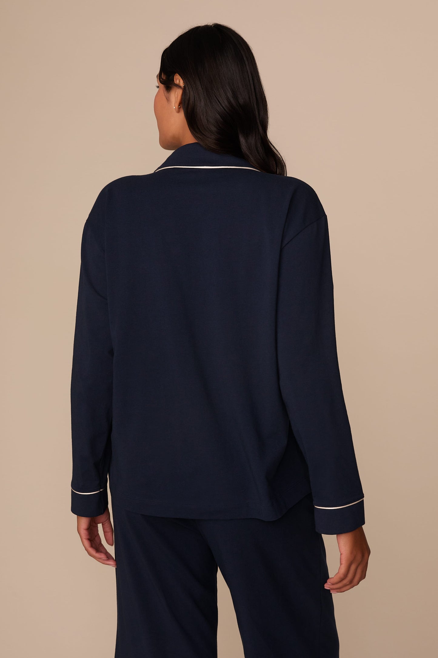 On a neutral studio backdrop, Victoria wears the Perfect Pima Pajama Long Sleeve Shirt in Navy with matching pajama pants. The back view highlights the relaxed silhouette, long sleeves, and contrast piping along the collar and cuffs.