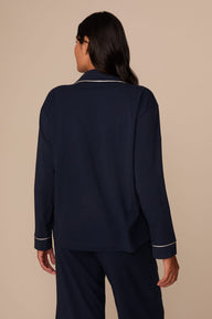 On a neutral studio backdrop, Victoria wears the Perfect Pima Pajama Long Sleeve Shirt in Navy with matching pajama pants. The back view highlights the relaxed silhouette, long sleeves, and contrast piping along the collar and cuffs.
