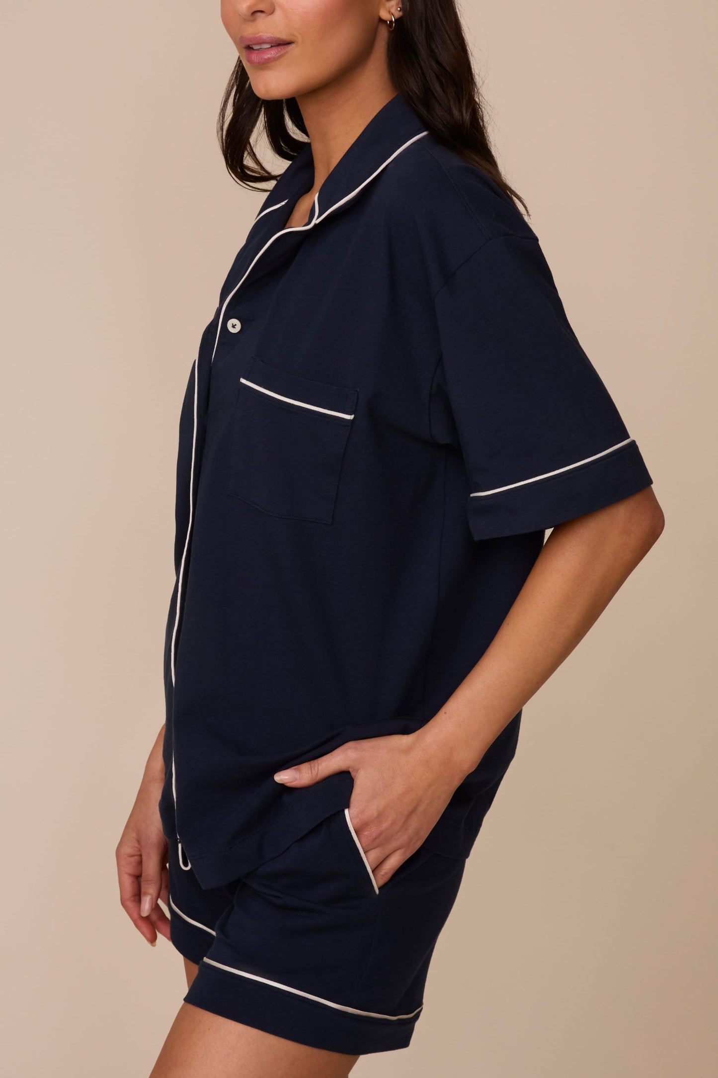 On a neutral studio backdrop, Marcela wears the Perfect Pima Pajama Shirt in Navy with matching navy pajama shorts. The side view highlights the relaxed silhouette, chest pocket, contrast piping details, and side pockets on the shorts.