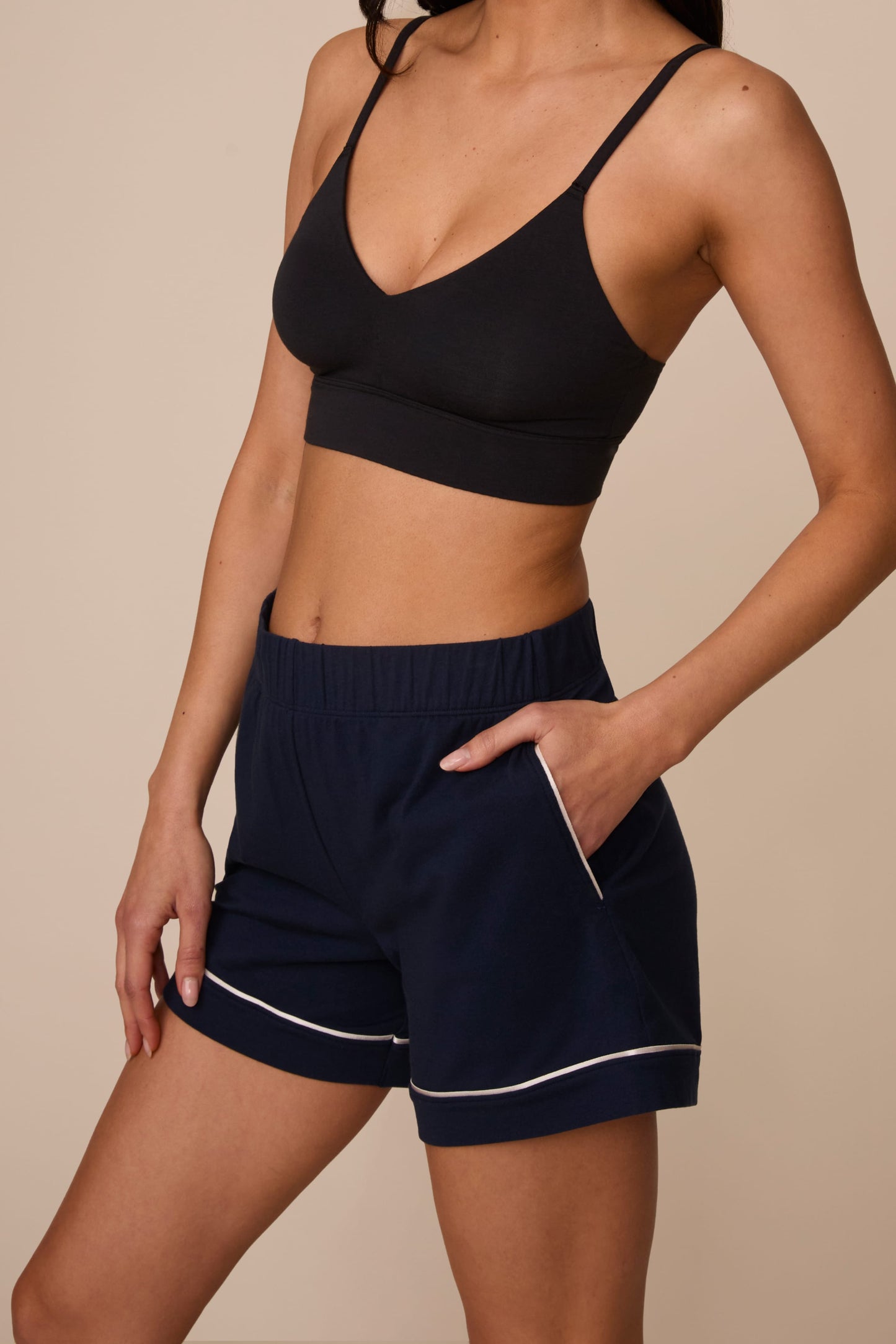 On a neutral studio backdrop, Marcela wears the Perfect Pima Pajama Short in Navy with a black bralette. The angled close-up highlights the elastic waistband, side pocket, relaxed fit, and contrast piping along the hem and pocket opening.