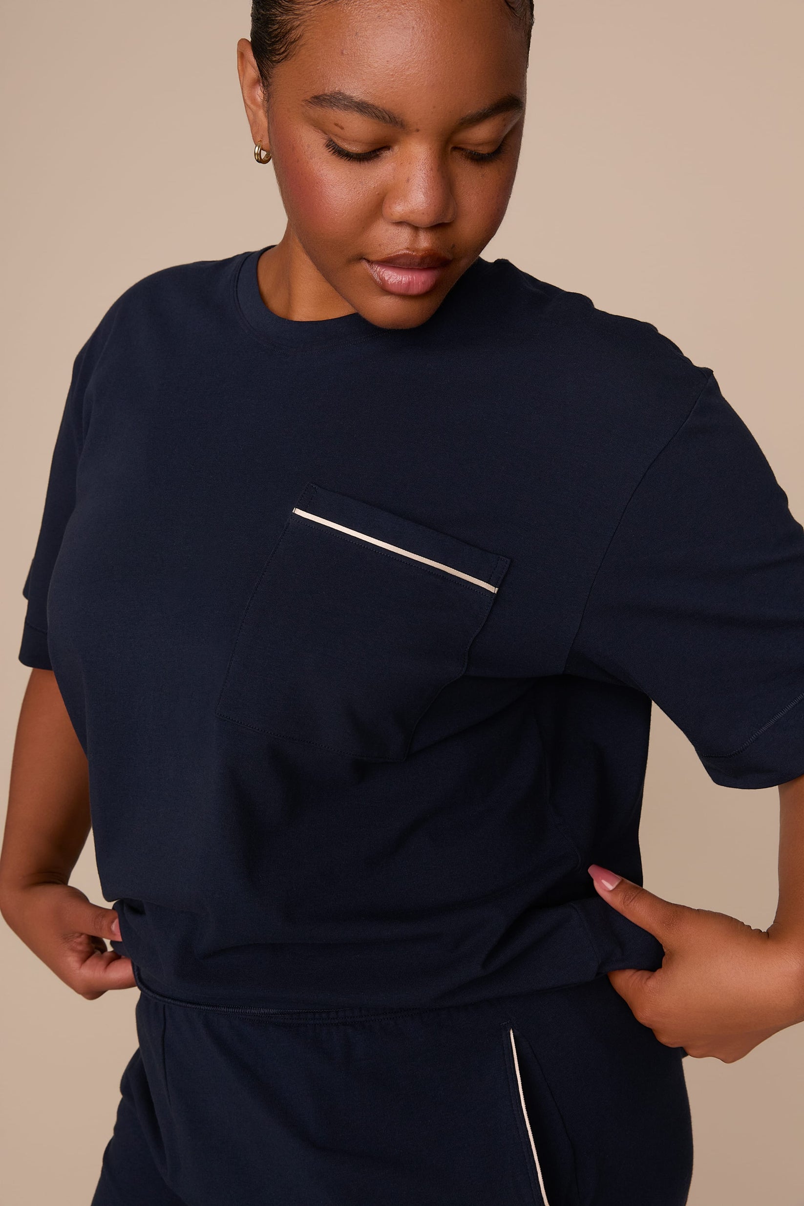 On a warm beige studio backdrop, Zarria wears the Perfect Pima Pajama T-Shirt in a deep navy color with matching pajama shorts. The close-up view highlights the relaxed fit, chest pocket with contrast piping, and soft drape of the fabric.