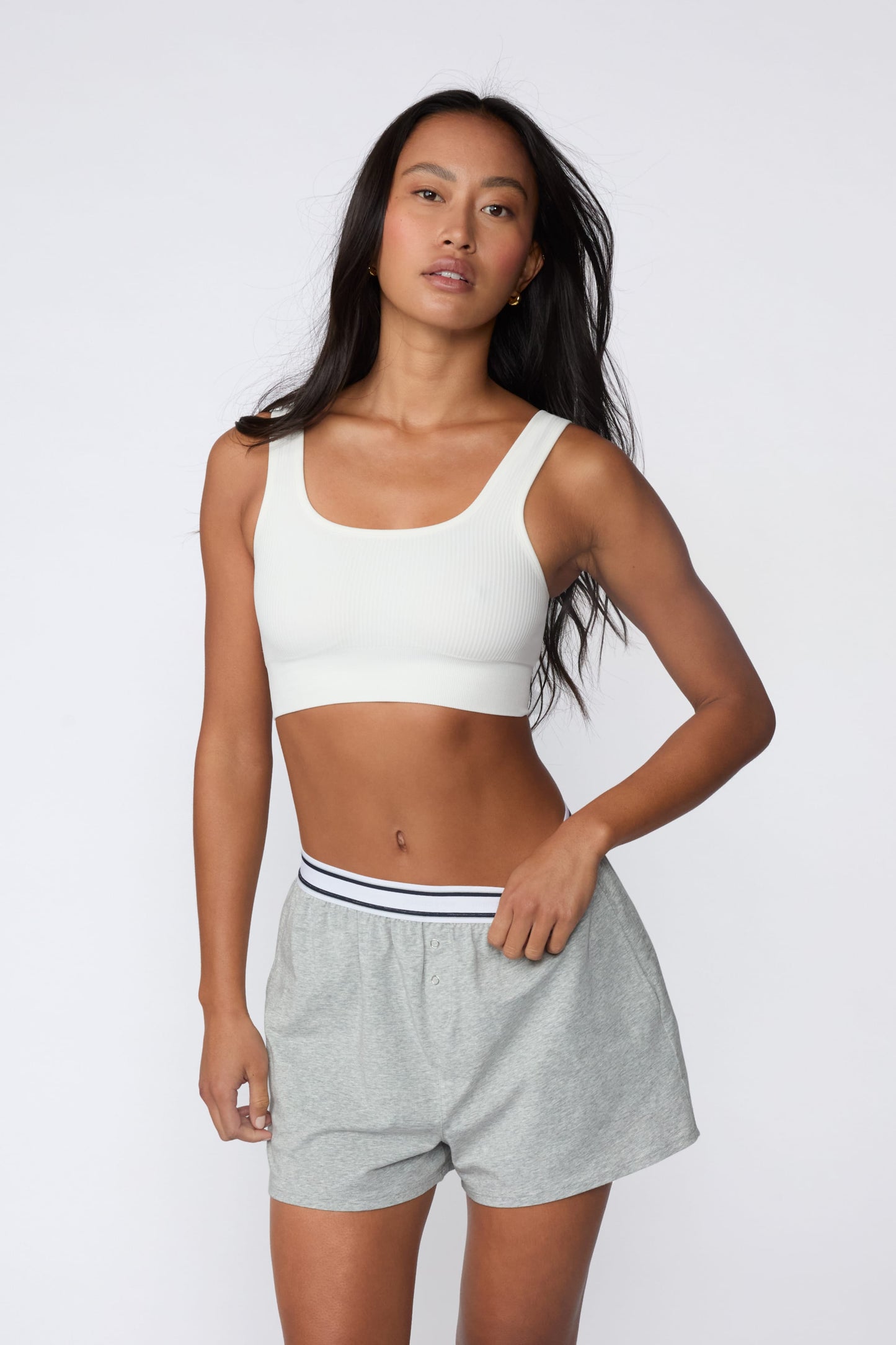 On a light gray studio backdrop, Amber wears the Ribbed Seamless Scoop Bralette in White with gray lounge shorts. A full-body view highlights the scoop neckline, wide straps, ribbed texture, and supportive underband.