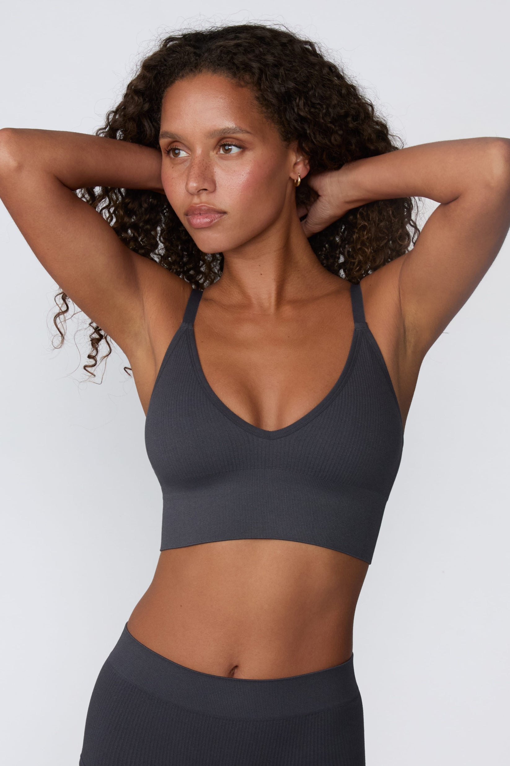 On a light gray studio backdrop, Courtney wears the Ribbed Seamless Triangle Bralette in Charcoal with a matching ribbed bottom. With her hands behind her head, she showcases the ribbed texture, V-neckline, adjustable straps, and wide underband.