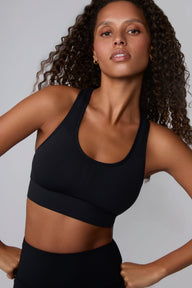 On a light gray studio backdrop, Courtney wears the Seamless Racerback Sports Bra in Black with matching bike shorts. The angled close-up highlights the scoop neckline, racerback straps, seamless construction, and wide underband.