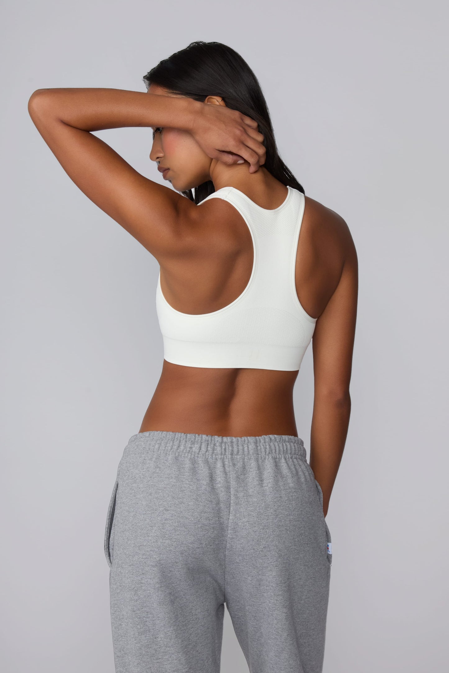 On a light gray studio backdrop, Amber wears the Seamless Racerback Sports Bra in White with gray sweatpants. The back view showcases the racerback design, breathable knit texture, and wide underband.