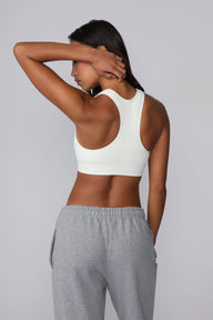 On a light gray studio backdrop, Amber wears the Seamless Racerback Sports Bra in White with gray sweatpants. The back view showcases the racerback design, breathable knit texture, and wide underband.