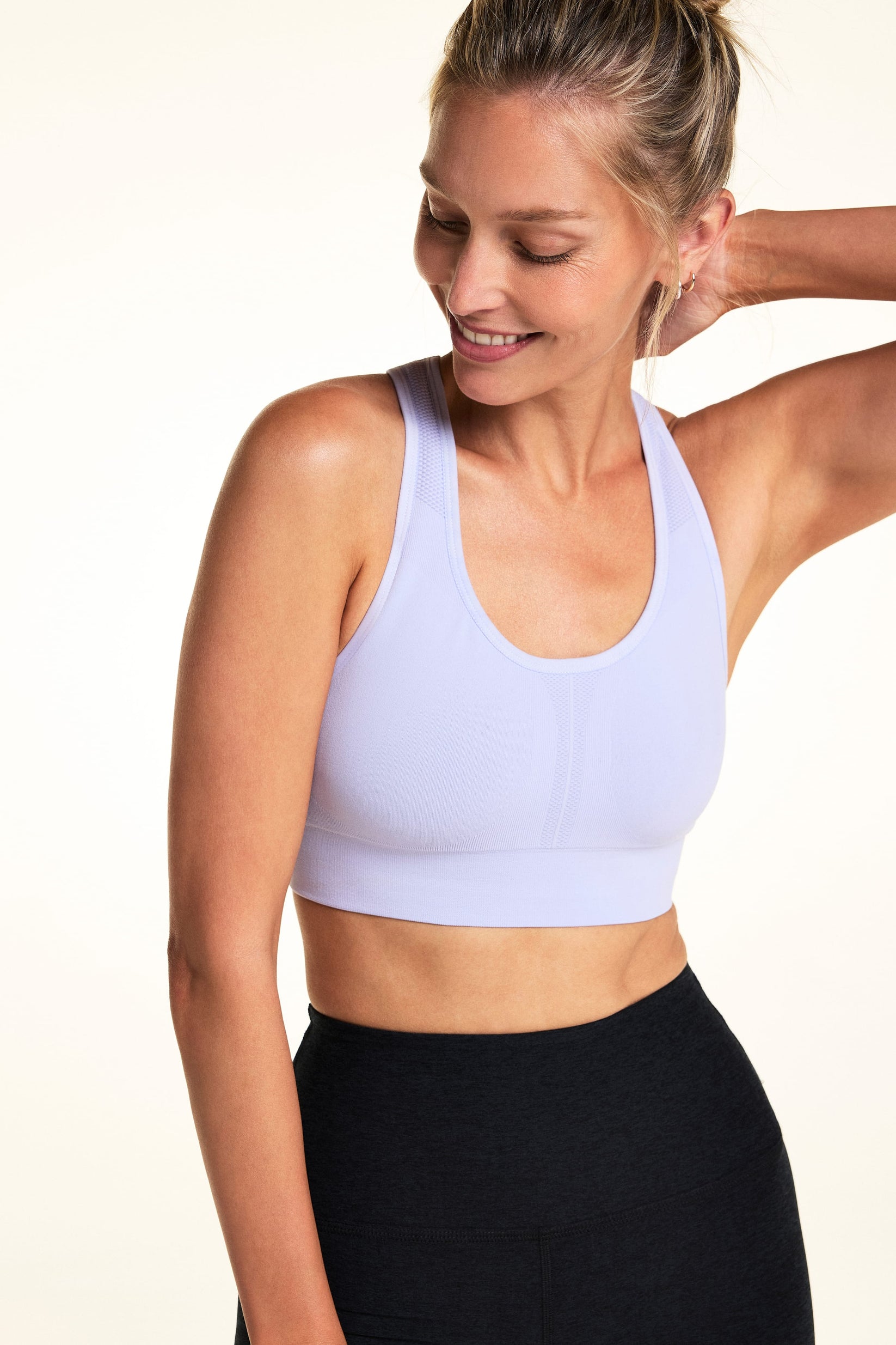 On a light studio backdrop, Meagan wears the Seamless Racerback Bra in Lilac with black high-rise bike shorts. The angled image highlights the scoop neckline, knit texture, and wide comfort band.