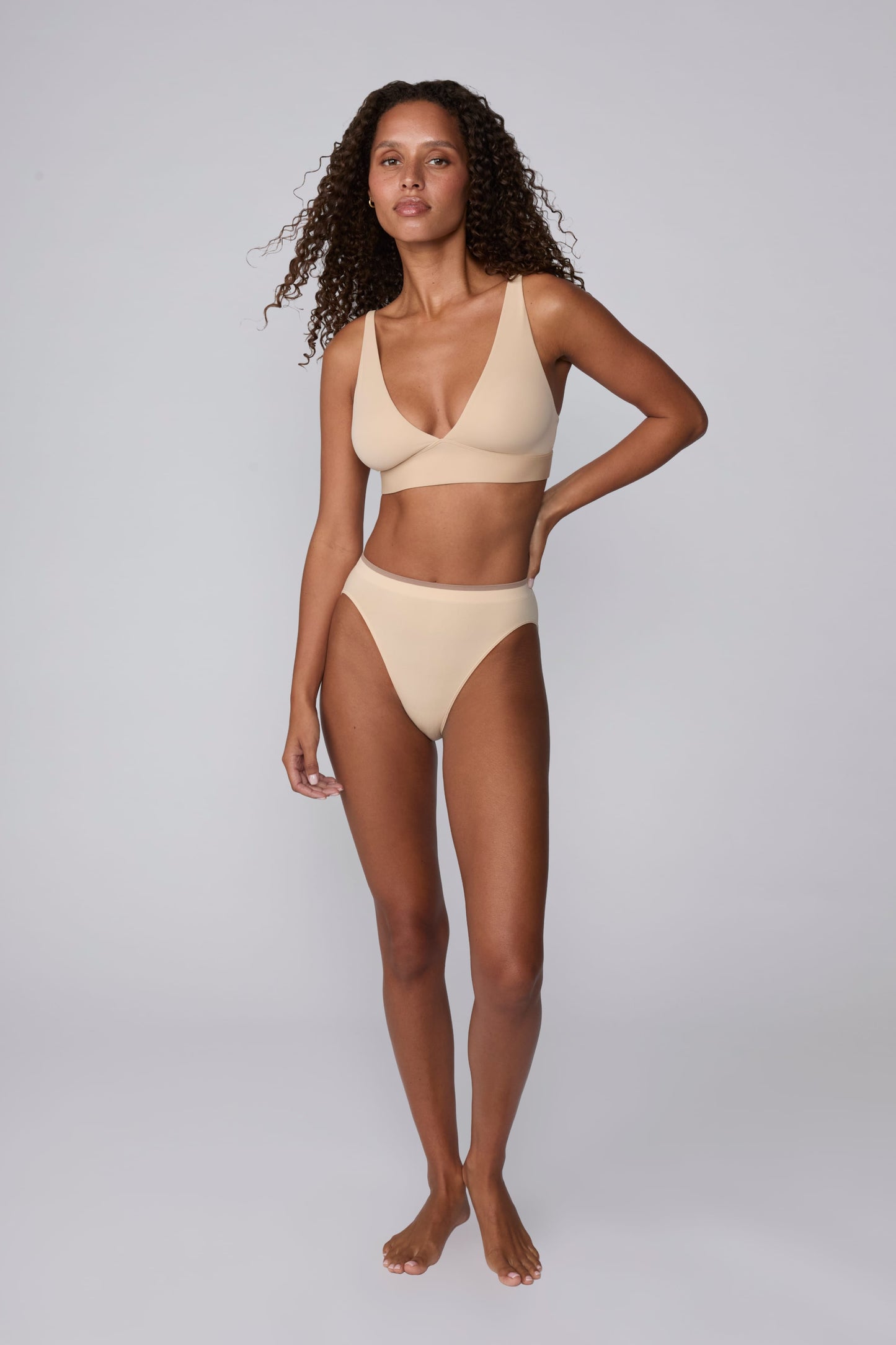 On a light gray studio backdrop, Courtney wears the Smooth Seamless Brief in Beige with the matching Smooth Seamless Bralette in Beige. The full-body front view highlights the high-leg fit, smooth finish, and contrast trim waistband.