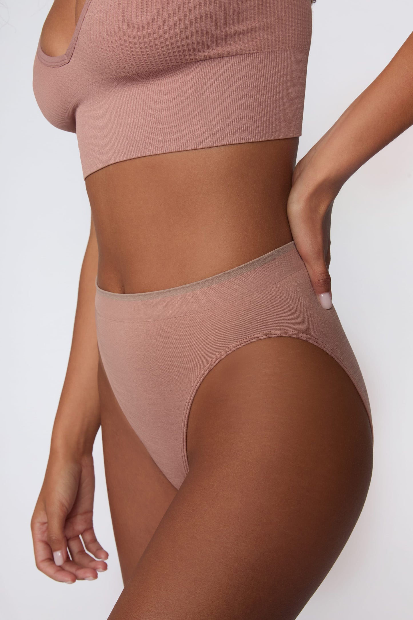 On a light gray studio backdrop, Courtney wears the Smooth Seamless Brief in Plum with the matching Seamless Scoop Bralette in Plum. A close-up angled view highlights the high-leg cut, smooth fabric, and contrast trim waistband.