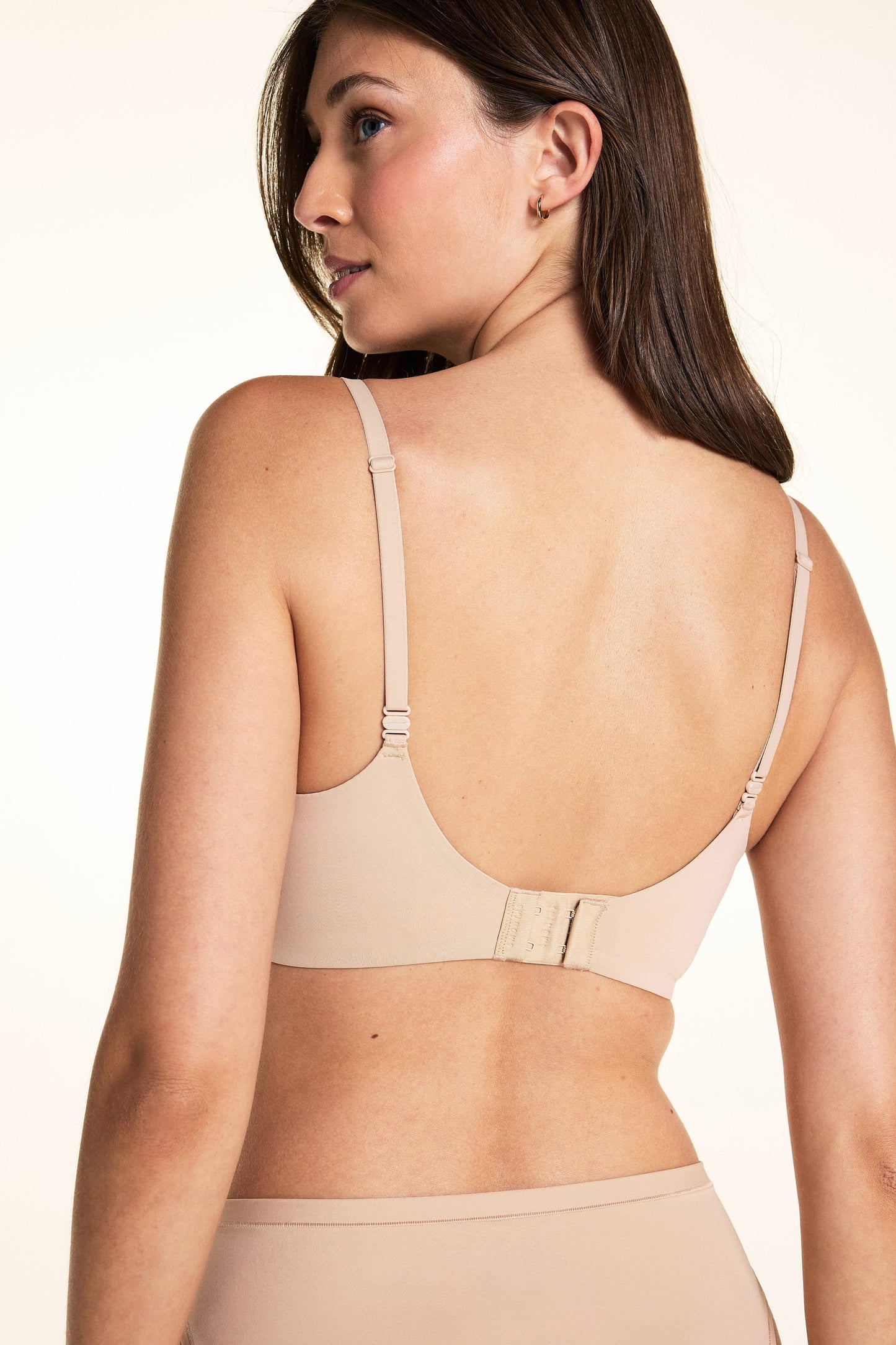On a light neutral backdrop, Laura is shown from the back wearing the Base Balconette Bra in Beige. The image highlights the adjustable straps, wide back band, and hook-and-eye closure.
