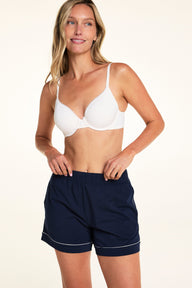 On a light studio backdrop, Meagan wears the Base Picot T-Shirt Bra in Ivory with navy lounge shorts. The angled front view highlights the smooth molded cups, picot trim, and adjustable straps.