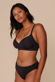 On a neutral beige backdrop, Victoria wears the Base Wirefree T-Shirt Bra in Black with matching underwear, highlighting the smooth molded cups, wireless design, and clean silhouette from an angled view.