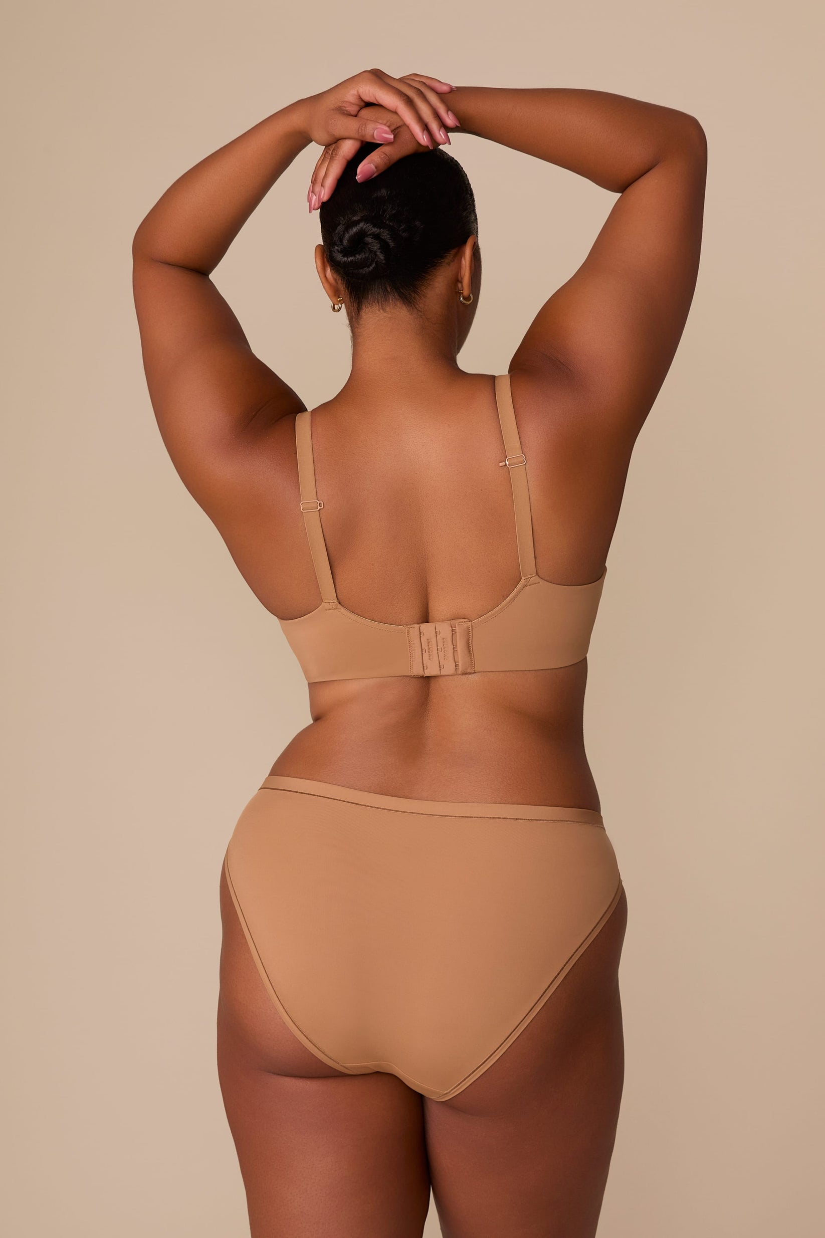 On a neutral beige backdrop, a back view of Zarria wearing the Base Wirefree T-Shirt Bra in Tan showcases the adjustable straps, hook-and-eye closure, and supportive band.