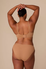 On a neutral beige backdrop, a back view of Zarria wearing the Base Wirefree T-Shirt Bra in Tan showcases the adjustable straps, hook-and-eye closure, and supportive band.