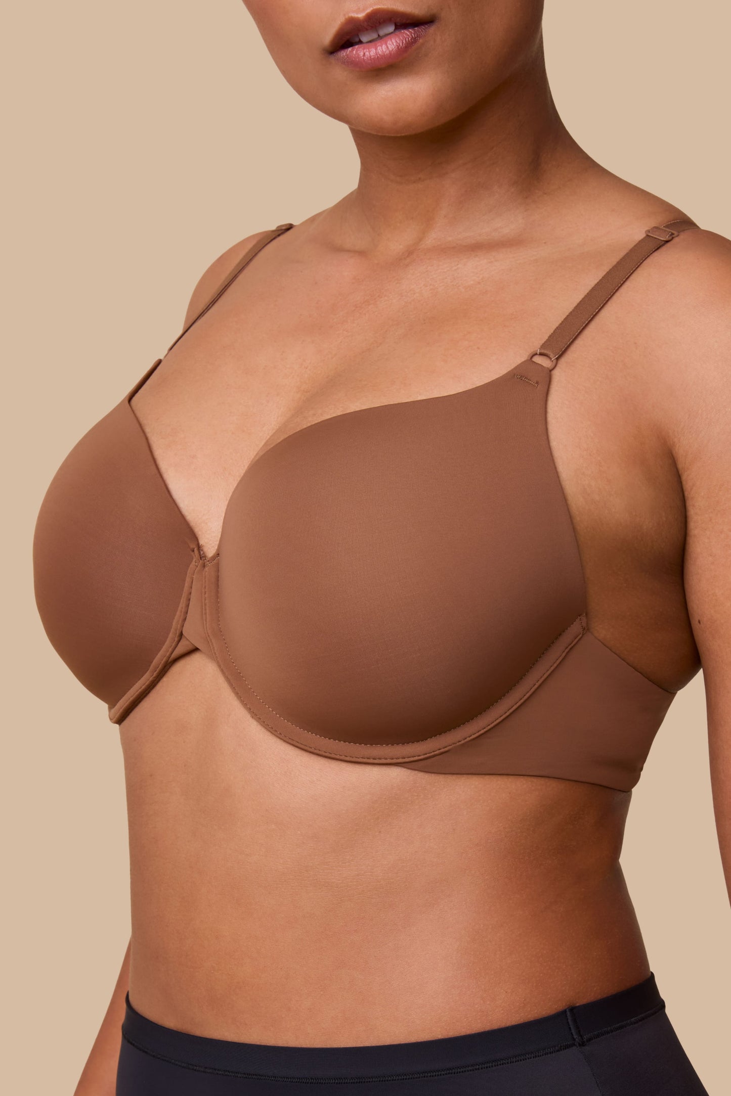 On a neutral beige backdrop, India wears the Base T-Shirt Bra in Brown, shown in a close-up view highlighting the lightly lined cups, signature X-bridge at the center front, and smooth microfiber fabric.